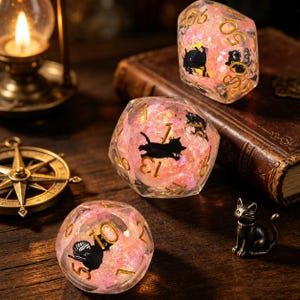 May include: Three polyhedral dice made of clear resin filled with pink iridescent glitter and small black cat silhouettes. Each die features gold-coloured numbers. The dice are arranged on a dark wooden surface alongside a small metal cat figurine.