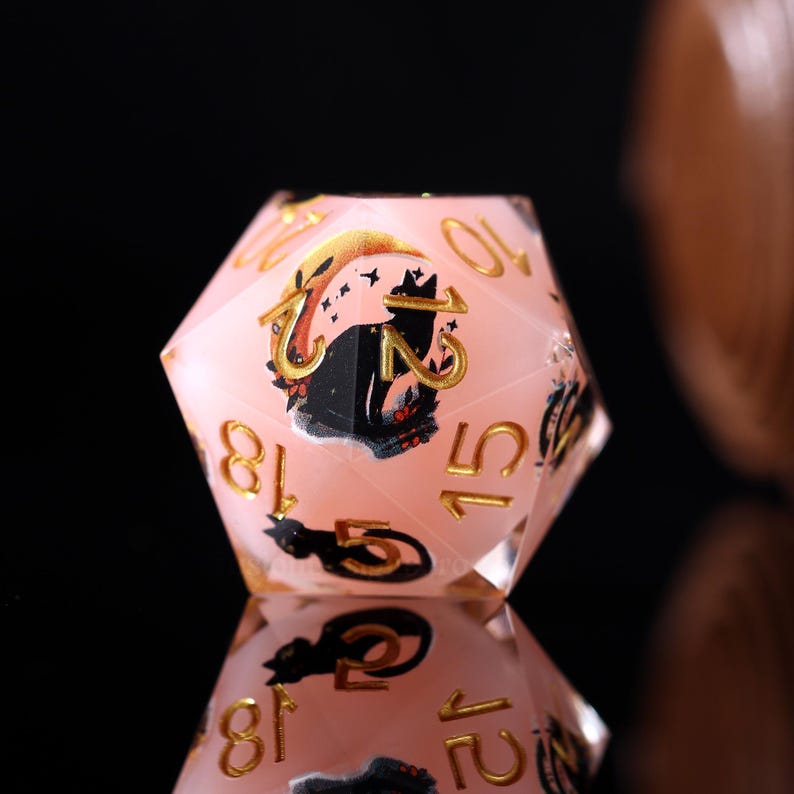 Pode incluir: Um dado rosa de vinte lados com numera&ccedil;&atilde;o dourada e ilustra&ccedil;&otilde;es de gatos pretos. O dado apresenta uma silhueta de gato preto com uma lua crescente e estrelas. Os n&uacute;meros est&atilde;o em uma fonte dourada estilizada.
