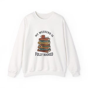 Op de afbeelding: Witte sweatshirt met de tekst "My Weekend Is Fully Booked". De afbeelding toont een stapel boeken, een kopje koffie en een bril. Een casual, comfortabele sweatshirt-stijl.
