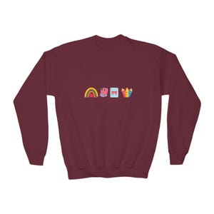 Op de afbeelding: Een bordeauxrode sweatshirt met een kleurrijke afbeelding van een regenboog, een roze cadeau, een blauw notitieboekje met een strik en een set kleurpotloden. Het ontwerp is gecentreerd op de borst. De sweatshirt heeft een ronde hals en lange mouwen.