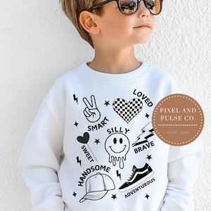 May include: White sweatshirt with black graphics including a peace sign, heart, lightning bolt, and the words "Smart", "Silly", "Loved", "Brave", "Sweet", "Handsome", and "Adventurous". A round logo says "Pixel and Pulse Co. ESTD 2025".