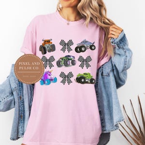 K&ouml;nnte beinhalten: Ein hellrosa T-Shirt mit bunten Monstertruck-Grafiken und karierten Schleifen. Die Trucks umfassen einen orangefarbenen Truck, einen Hai-Truck, einen gr&uuml;nen Truck, einen lila Einhorn-Truck und einen gr&uuml;nen Truck. Das Shirt hat auch den Text "PIXEL AND PULSE CO. ESTD 2023".