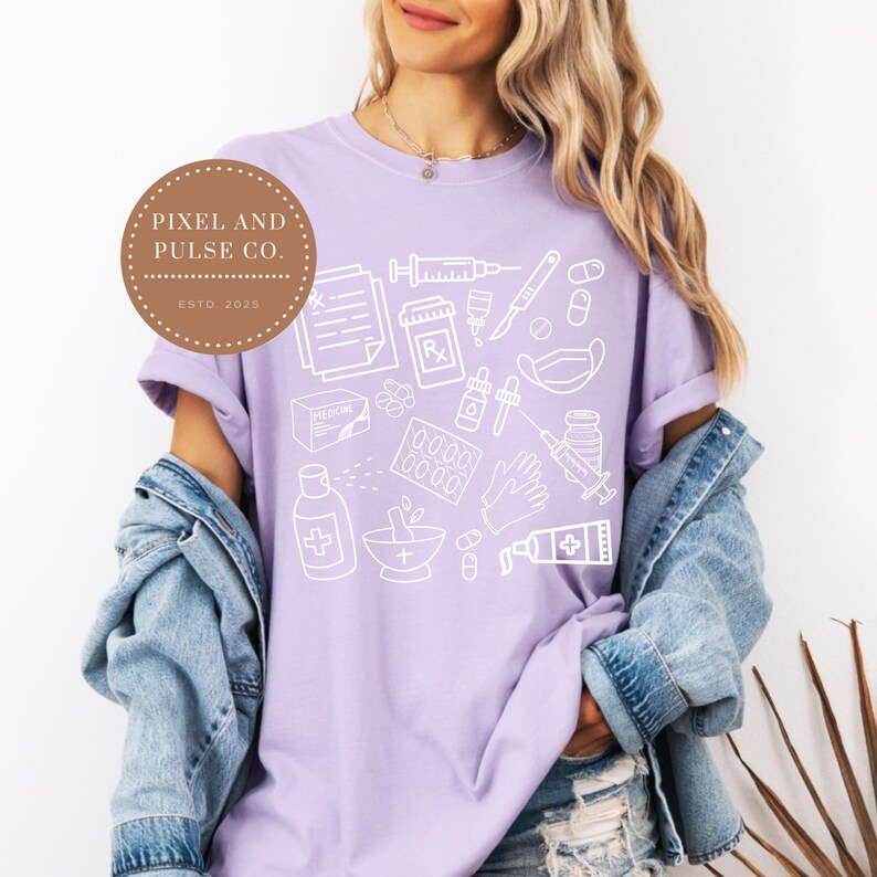 Puede incluir: Una camiseta lila con un gr&aacute;fico blanco de suministros m&eacute;dicos, incluyendo jeringas, pastillas y recetas. La camiseta tiene cuello redondo y mangas cortas. Un logotipo circular con el texto "Pixel and Pulse Co. ESTD 2020" es visible.
