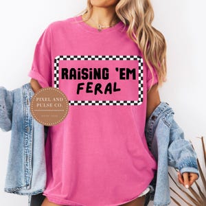 May include: A bright pink t-shirt with the text "RAISING 'EM FERAL" in a black and white checkered frame. The shirt also features a circular logo with the text "PIXEL AND PULSE CO. ESTD 2023".