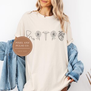 May include: Cream-colored t-shirt with a black line drawing of a stethoscope, sunflowers, a caduceus, and a human heart. The shirt also features a circular logo with the text "PIXEL AND PULSE CO. ESTD 2025".