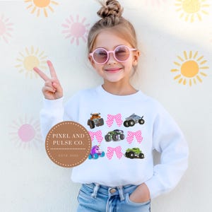 Op de afbeelding: Witte sweatshirt met kleurrijke monstertrucks en roze strikken. Het ontwerp bevat een oranje, grijze, groene en paarse monstertruck, evenals een eenhoorn-truck. De sweatshirt wordt gedragen door een jong persoon met een roze zonnebril.