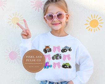 Könnte beinhalten: Weißes Sweatshirt mit bunten Monstertrucks und rosa Schleifen. Das Design umfasst einen orangefarbenen, grauen, grünen und lila Monstertruck sowie einen Einhorn-Truck. Das Sweatshirt wird von einer jungen Person mit rosa Sonnenbrille getragen.