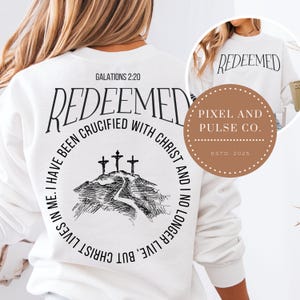 May include: White sweatshirt with the word "REDEEMED" printed in black. The back features a graphic of three crosses on a hill, with text from Galatians 2:20. The sweatshirt also has the text "I HAVE BEEN CRUCIFIED WITH CHRIST AND I NO LONGER LIVE, BUT CHRIST LIVES IN ME."