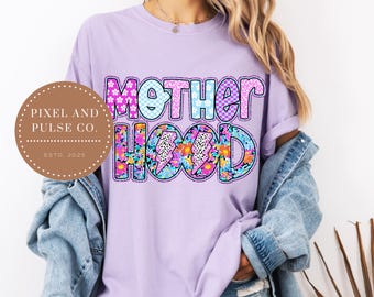 May include: A light purple t-shirt with the word "MOTHERHOOD" in a colorful, patterned design. The word "MOTHER" is in a floral and checkered pattern, and "HOOD" features a floral design with lightning bolts. The shirt also has a Pixel and Pulse Co. logo.