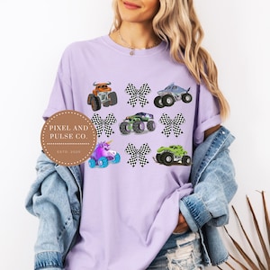 K&ouml;nnte beinhalten: Ein helllila T-Shirt mit bunten Monster-Truck-Designs und karierten Schleifen. Die Trucks sind in verschiedenen Farben, darunter Orange, Gr&uuml;n und Blau. Das T-Shirt hat auch den Text "Pixel and Pulse Co. Est. 2023".