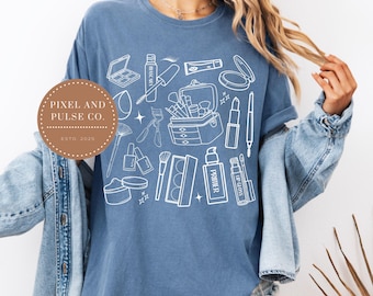 May include: A blue t-shirt featuring a white outline design of various makeup items, including a makeup case, lipstick, mascara, and brushes. The shirt also has the text "PIXEL AND PULSE CO. ESTD. 2025" in a circular logo.