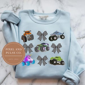 K&ouml;nnte beinhalten: Hellblaues Sweatshirt mit bunten Monstertruck-Designs, darunter ein Hai, ein Einhorn und ein Stier. Das Design enth&auml;lt auch karierte Schleifen. Das Sweatshirt hat den Text "PIXEL AND PULSE CO. ESTD. 2025" in einem kreisf&ouml;rmigen Logo.