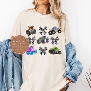 K&ouml;nnte beinhalten: Cremefarbenes T-Shirt mit farbenfrohen Monstertruck-Grafiken und karierten Schleifen. Das Design umfasst einen orangefarbenen Truck, einen Hai-Truck, einen gr&uuml;nen Truck, einen lila Einhorn-Truck und mehr. Das Shirt tr&auml;gt auch den Text "PIXEL AND PULSE CO. ESTD 2023".