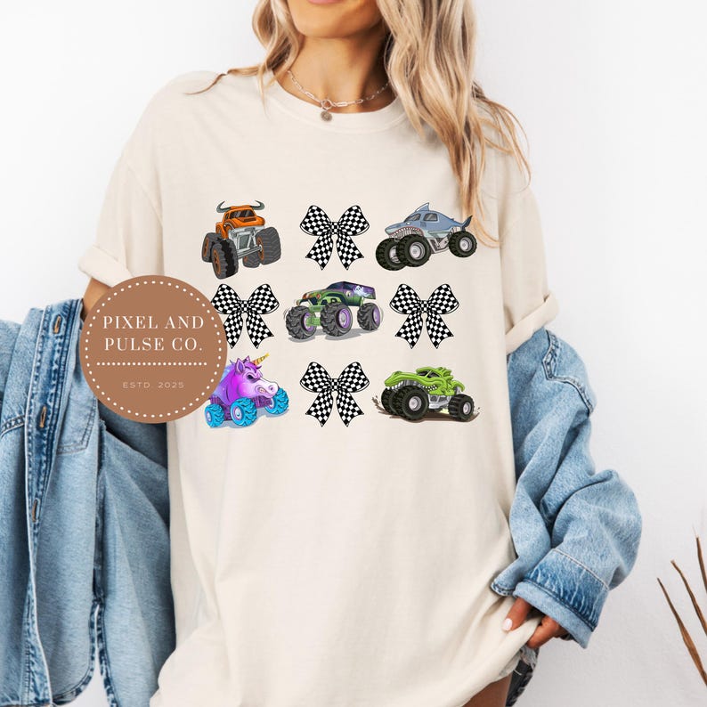 K&ouml;nnte beinhalten: Cremefarbenes T-Shirt mit farbenfrohen Monstertruck-Grafiken und karierten Schleifen. Das Design umfasst einen orangefarbenen Truck, einen Hai-Truck, einen gr&uuml;nen Truck, einen lila Einhorn-Truck und mehr. Das Shirt tr&auml;gt auch den Text "PIXEL AND PULSE CO. ESTD 2023".