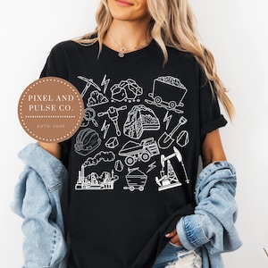 Puede incluir: Camiseta negra con una ilustración en blanco de contornos de herramientas, equipos y estructuras mineras. El diseño incluye un pico, rocas, un vagón de carbón, un casco y un pozo petrolífero. La camiseta también presenta el texto "PIXEL AND PULSE CO. ESTD 2025".