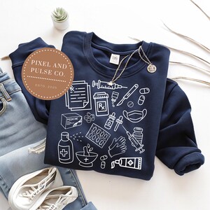 Puede incluir: Sudadera azul marino con ilustraciones m&eacute;dicas blancas, incluyendo jeringas, pastillas y s&iacute;mbolos de receta. La sudadera se combina con vaqueros azules y zapatillas blancas. Se ve un collar con un colgante.