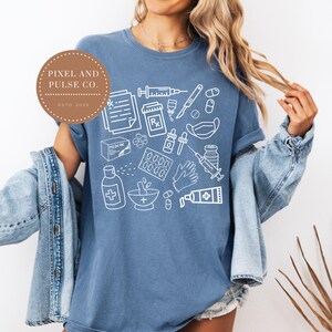 Puede incluir: Una camiseta azul con un gr&aacute;fico blanco de suministros m&eacute;dicos, incluyendo jeringas, pastillas y s&iacute;mbolos de recetas. La camiseta tiene un logotipo redondo marr&oacute;n que dice "PIXEL AND PULSE CO. ESTD. 2023".