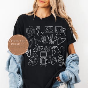 May include: Black t-shirt featuring white line drawings of medical imagery, including a heart, ultrasound scans, and a kidney. The shirt also has the text "PIXEL AND PULSE CO. ESTD. 2025" on a circular label. A medical-themed design.