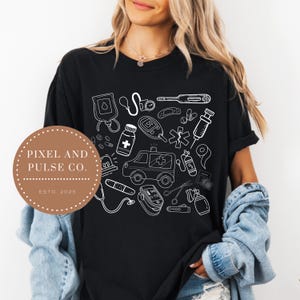 May include: Black t-shirt featuring a white medical-themed graphic. The design includes an ambulance, medical tools, pills, and a blood bag. The shirt also has the text "Pixel and Pulse Co. Estd 2025" on a circular label.