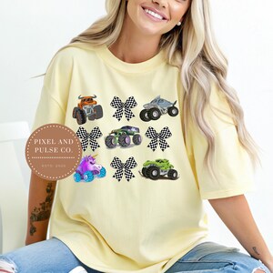 K&ouml;nnte beinhalten: Ein hellgelbes T-Shirt mit farbenfrohen Monster-Truck-Grafiken und karierten Schleifen. Die Trucks umfassen einen orangefarbenen Truck, einen Hai-Truck, einen gr&uuml;nen Truck, einen lila Einhorn-Truck und mehr. Das Shirt hat auch den Text "Pixel and Pulse Co. Estd. 2023".