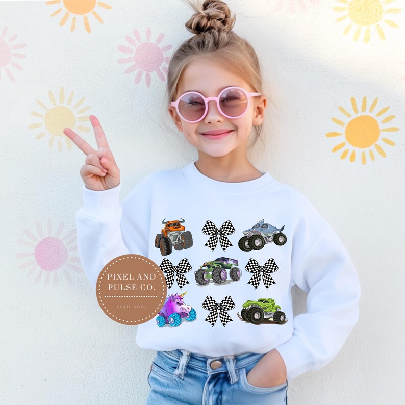 K&ouml;nnte beinhalten: Wei&szlig;es Sweatshirt mit farbenfrohen Monstertruck-Grafiken und karierten Schleifen. Das Design umfasst orange, gr&uuml;ne, lila und graue Trucks. Das Sweatshirt wird von einer jungen Person mit rosa Sonnenbrille getragen.