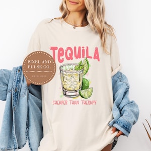 May include: Cream-colored t-shirt with the word "Tequila" in pink script above a graphic of a tequila shot with lime and mint. The phrase "Cheaper Than Therapy" is below the shot. A circular logo is on the left.