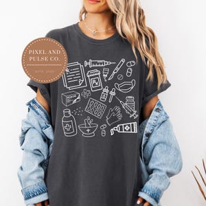 Puede incluir: Camiseta gris oscuro con un dise&ntilde;o de contorno blanco de suministros m&eacute;dicos, incluyendo jeringas, pastillas y s&iacute;mbolos de receta. La camiseta tambi&eacute;n tiene el texto "Pixel and Pulse Co. ESTD 2025" en un dise&ntilde;o circular. Se lleva una chaqueta vaquera.