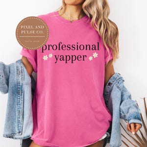 May include: A bright pink t-shirt with the words "professional yapper" in black script, accented by small white flower graphics. The shirt is part of a casual outfit, paired with a denim jacket.
