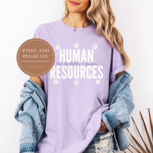 May include: A light purple t-shirt with the words "HUMAN RESOURCES" in white, with the text "dept. for children & families" underneath. The shirt also features small white daisy graphics. A brown circular logo with the text "PIXEL AND PULSE CO. ESTD 2025" is on the left.