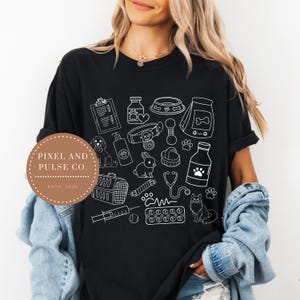 May include: Black t-shirt featuring a white outline design of pet care items, including a dog, cat, bunny, food bowl, medicine, and a stethoscope. The shirt also has the text "Pixel and Pulse Co. Estd. 2025" in a circular design.