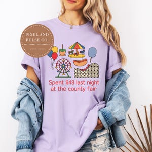 Puede incluir: Camiseta lila con un diseño colorido de feria. El diseño incluye una noria, una montaña rusa, un tiovivo, un perrito caliente y globos. El texto de la camiseta dice: "Spent 44€ last night at the county fair."