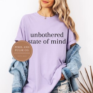 May include: A light purple t-shirt with the words "unbothered state of mind" in black. A brown circular logo with the text "Pixel and Pulse Co. Estd. 2025" is on the left. The model is wearing a denim jacket.