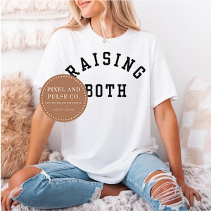 Könnte beinhalten: Weißes T-Shirt mit dem schwarzen Text "RAISING BOTH", der sich über dem Wort "BOTH" wölbt. Ein kreisförmiges Logo mit dem Text "PIXEL AND PULSE CO." und "ESTD. 2025" befindet sich links. Die Person trägt zerrissene blaue Jeans.