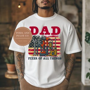 May include: White t-shirt with a graphic featuring the word "DAD" in red above an American flag design with tools. The phrase "FIXER OF ALL THINGS" is printed below the flag. A circular logo is on the left side.