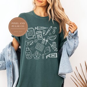 Puede incluir: Camiseta verde oscuro con dibujo lineal blanco de suministros m&eacute;dicos, incluyendo jeringas, pastillas y recetas. La camiseta tambi&eacute;n presenta el texto "PIXEL AND PULSE CO. ESTD 2025" en un dise&ntilde;o circular.