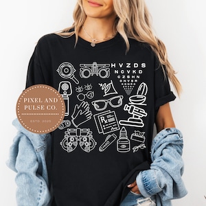 Könnte beinhalten: Schwarzes T-Shirt mit weißer Strichzeichnung von Augenpflege-Werkzeugen, einer Sehtafel und dem Text "PIXEL AND PULSE CO. ESTD 2025". Das Design umfasst Brillen, Augentropfen und andere Illustrationen zur Augenheilkunde.