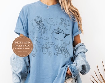 May include: Light blue t-shirt featuring a detailed black line drawing of various sea creatures, including a jellyfish, stingray, turtle, dolphin, whale, and octopus. The shirt has a relaxed fit. A circular logo with the text "PIXEL AND PULSE CO. ESTD. 2023" is on the left.