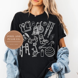 Può includere: T-shirt nero con illustrazioni bianche di attrezzature mediche e fisioterapiche, tra cui una sedia a rotelle, stampelle e una palla da ginnastica. La maglietta presenta anche un logo che dice "PIXEL AND PULSE CO. ESTD. 2025."