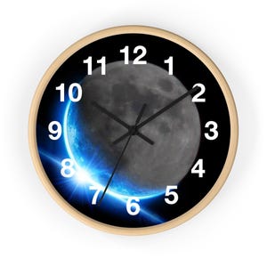 Puede incluir: Un reloj de pared redondo con fondo negro y marco de madera blanco. La esfera del reloj presenta una luna creciente con un brillo azul y un fondo negro. Los números del 1 al 12 están impresos en blanco alrededor de la esfera del reloj.