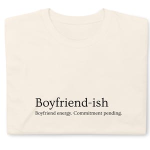 Puede incluir: Camiseta crema doblada con el texto negro "Boyfriend-ish" y la frase "Boyfriend energy. Commitment pending." Una camiseta sencilla e informal, perfecta para el uso diario.