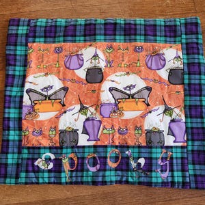 May include: A teal and purple plaid fabric with a Halloween-themed print featuring witches, cats, and cauldrons. The fabric has the word "SPOOKY" written in a whimsical font.