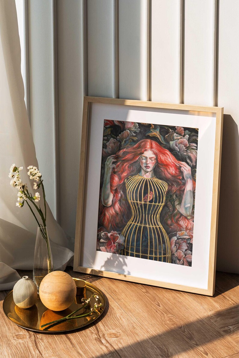Op de afbeelding: Een ingelijste kunstdruk met een schilderij van een persoon met lang, golvend rood haar, gekleed in een geel kooivormig lijfje over een donker kledingstuk, omringd door donkere bloemelementen. Het kunstwerk zit in een licht houten lijst.