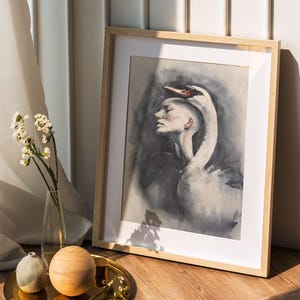 May include: A framed art print featuring a watercolor painting of a person with a shaved head and a white swan draped over their shoulders. The artwork uses shades of gray, black, and white with a red accent on the swan's beak, set in a light wood frame.