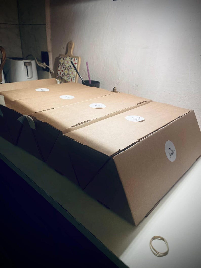 May include: Three rectangular brown cardboard boxes arranged in a row. Each box features a smooth, matte finish and a white circular sticker on the top and side surfaces. The boxes are constructed from folded paperboard with clean, sharp edges.