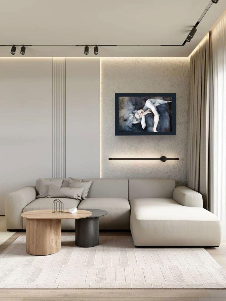 May include: A rectangular framed wall art piece featuring a monochromatic illustration of a reclining figure in shades of black, gray, and white. The artwork is displayed on a textured wall above a beige sectional sofa and two round coffee tables.