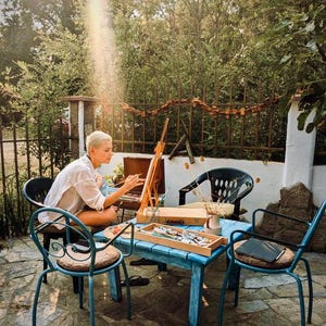 May include: A person with short blonde hair wearing a white shirt sits at a blue wooden table painting on a wooden easel. The table holds a wooden paint box, brushes, and a small ceramic vase with dried stems. Several metal chairs surround the table.