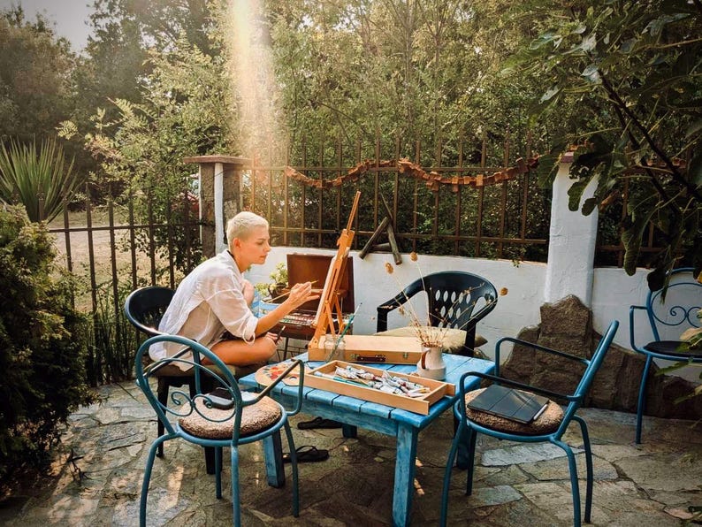 May include: A person with short blonde hair wearing a white shirt sits at a blue wooden table painting on a wooden easel. The table holds a wooden paint box, brushes, and a small ceramic vase with dried stems. Several metal chairs surround the table.