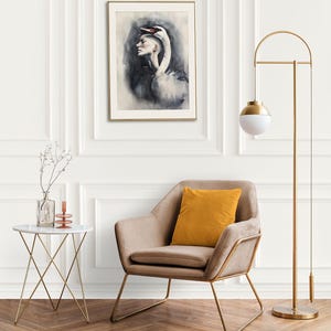 May include: A framed wall art print featuring a surreal illustration of a person with a swan head, displayed in a room with a beige armchair, a mustard yellow pillow, a white side table, and a tall gold floor lamp with a white globe shade.