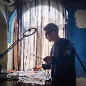 May include: A person with short hair wearing a dark long-sleeved shirt, standing at a table while painting on paper with a brush. A circular ring light on an adjustable stand illuminates the workspace, and a smartphone is mounted on a tripod nearby.