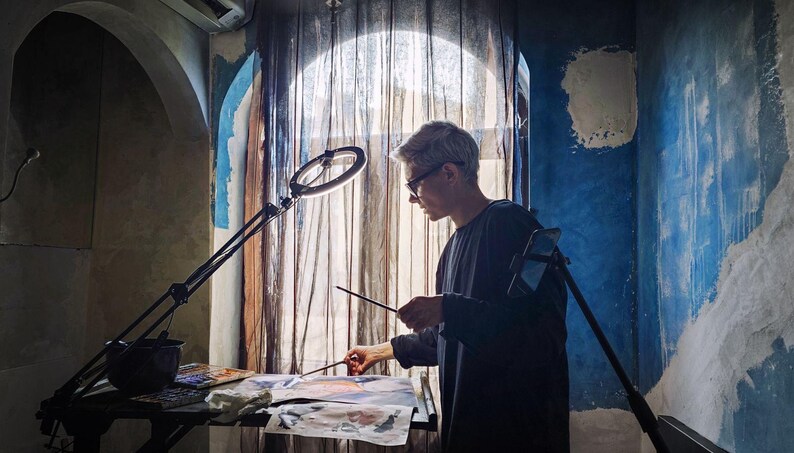 May include: A person with short hair wearing a dark long-sleeved shirt, standing at a table while painting on paper with a brush. A circular ring light on an adjustable stand illuminates the workspace, and a smartphone is mounted on a tripod nearby.